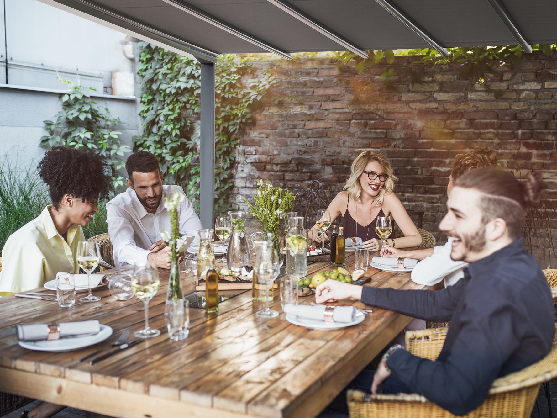 [Translate to Französisch:] Freunde sitzen gemeinsam unter einer Pergola am Abendbrottisch