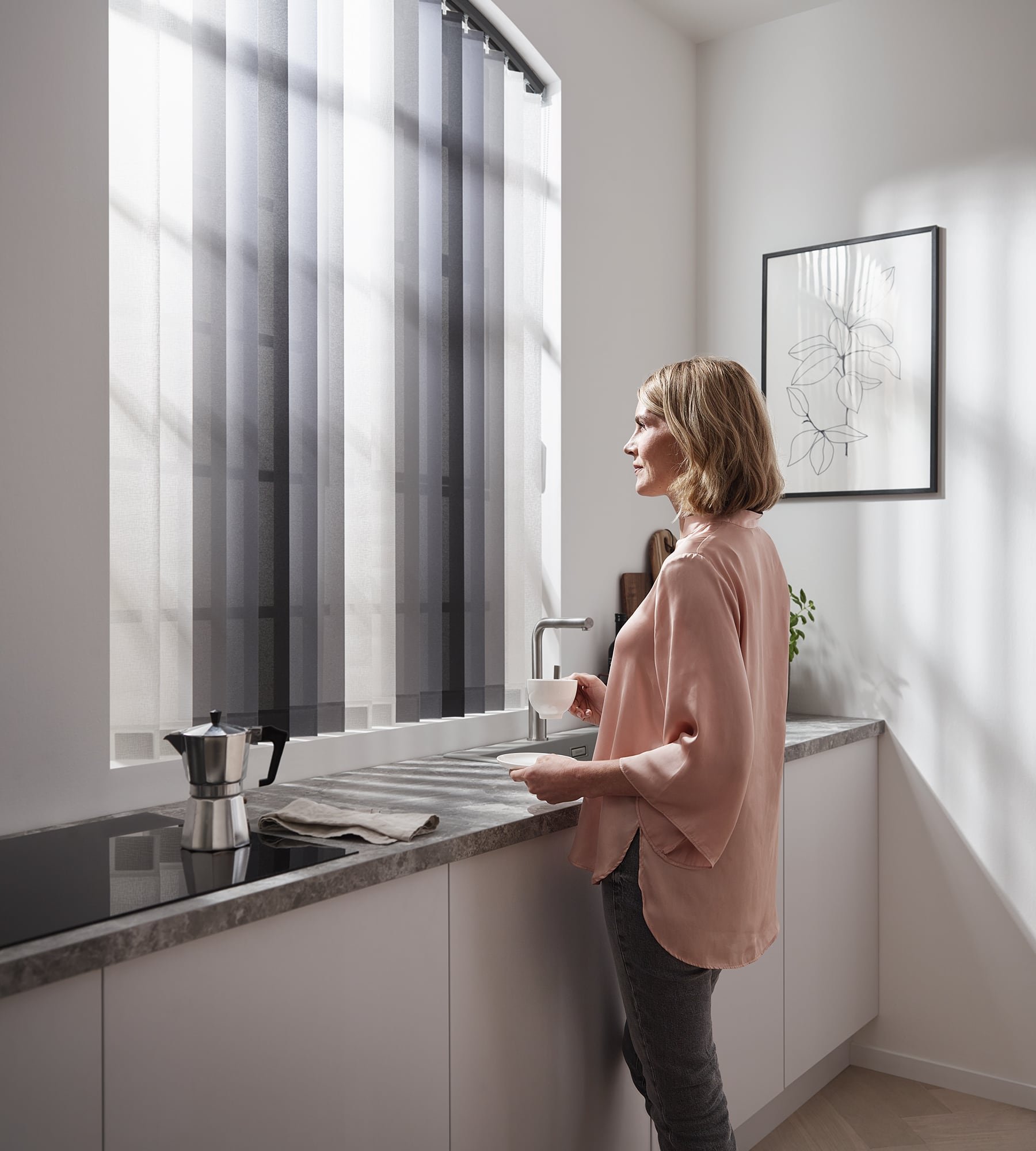 Frau sieht aus Bogenfenster mit KADECO Lamellenvorhang mit Farbwechsel in der Küche